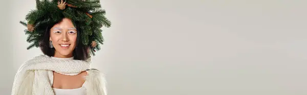Зимнее знамя, счастливая азиатка с натуральным сосновым венком, позирующим в белой одежде на сером фоне — стоковое фото