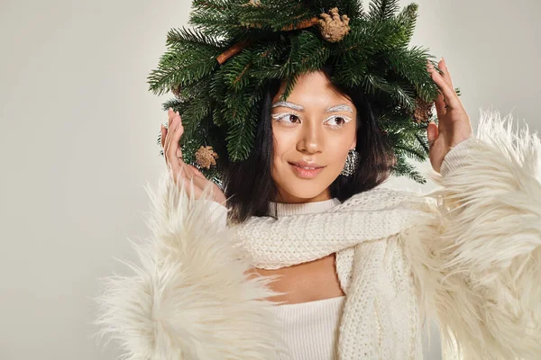 Belleza de invierno, mujer positiva con corona de pino natural posando en ropa blanca sobre fondo gris - foto de stock