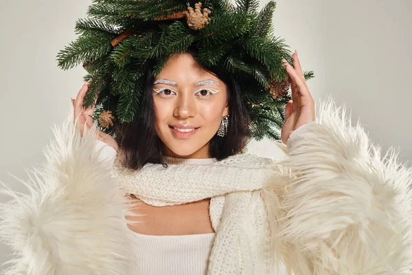 Зимняя красота, радостная азиатская женщина с натуральным сосновым венком позирует в белой одежде на сером фоне — стоковое фото