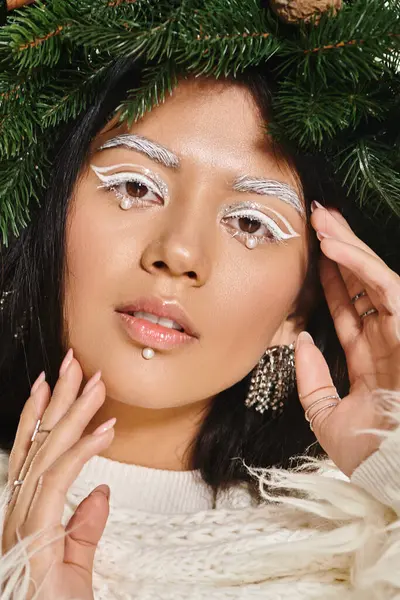 Winter trends, asian woman with white eye makeup and beads on face posing in wreath, look at camera — Stock Photo