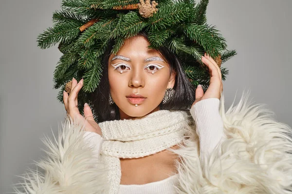 Winter trends, attractive woman with white eye makeup and beads on face posing in wreath on grey — Stock Photo