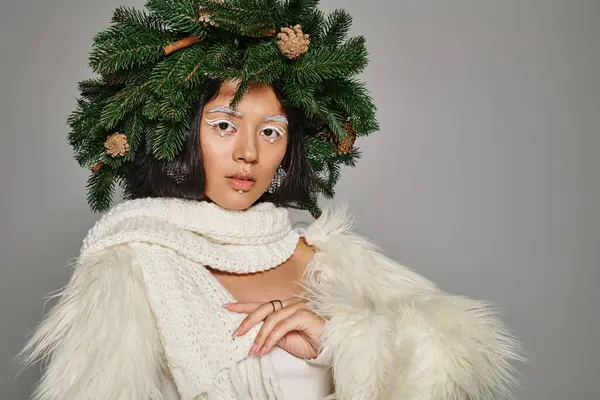 Праздничный стиль, азиатская зимняя королева с белым макияжем глаз и бусы на лице позируя в венке на сером — стоковое фото