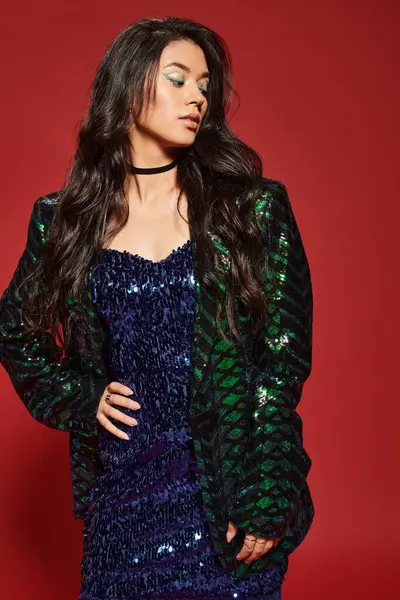 Brunette asian woman in green dress and jacket with sequins posing with hand on hip on red backdrop — Stock Photo