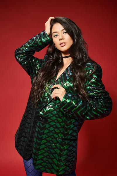Asian brunette young woman in trendy green jacket with sequins looking at camera on red backdrop — Stock Photo