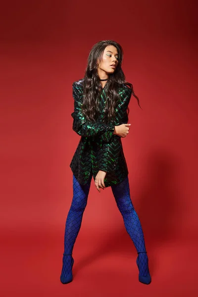 Mulher atraente na moda casaco verde com lantejoulas e meia-calça azul posando no fundo vermelho — Fotografia de Stock