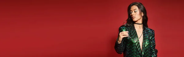 Брюнетка азиатская женщина в зеленой куртке с блестками держа очки на красном фоне, баннер — стоковое фото