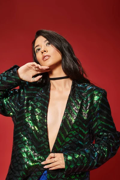 Mujer asiática soñadora en chaqueta verde de moda con lentejuelas mirando hacia arriba y posando sobre fondo rojo - foto de stock