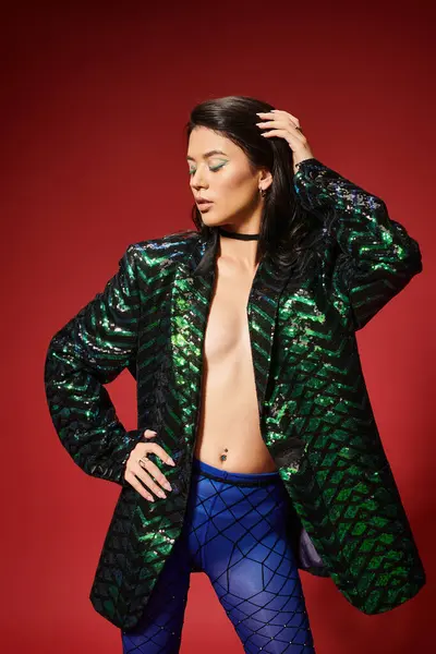Sexy asian woman in trendy green jacket with sequins posing with hand on hip on red backdrop — Stock Photo