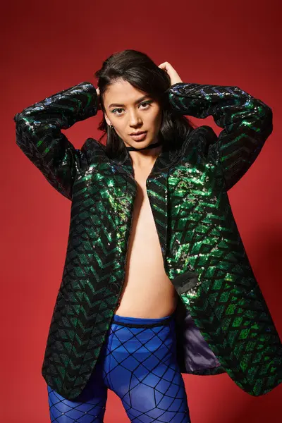 Jovem asiático modelo na moda jaqueta verde com lantejoulas posando com as mãos perto do cabelo no fundo vermelho — Fotografia de Stock