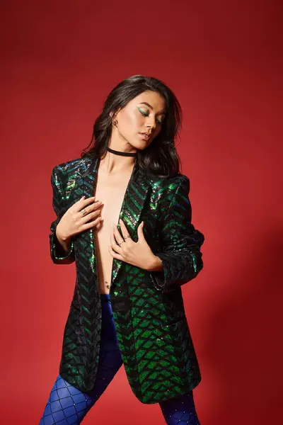 Sexy asian woman in stylish green jacket with sequins posing with hand on hip on red backdrop — Stock Photo
