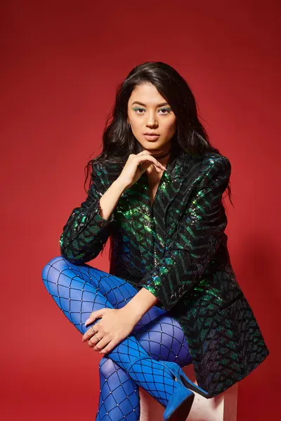 Pretty asian woman in stylish green jacket with sequins and blue pantyhose sitting on red backdrop — Stock Photo