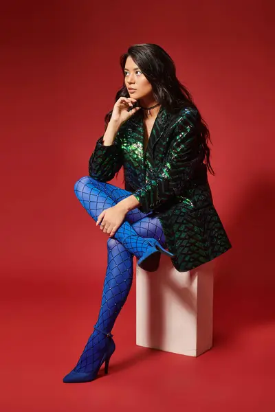 Stylish asian woman in stylish green jacket with sequins and blue pantyhose sitting on red backdrop — Stock Photo