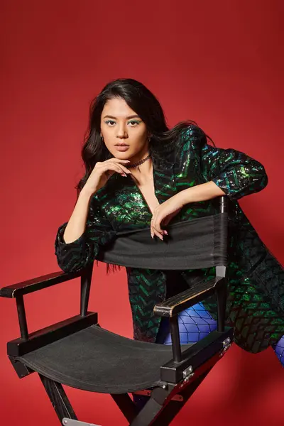 Young asian woman in green jacket with sequins and blue pantyhose posing near chair on red backdrop — Stock Photo