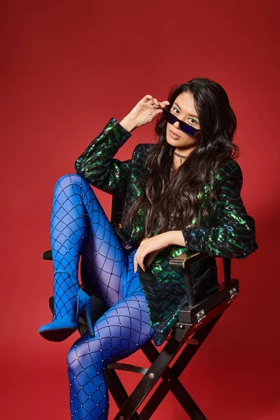 Stylish asian woman in sunglasses and green jacket with sequins sitting on chair on red backdrop — Stock Photo