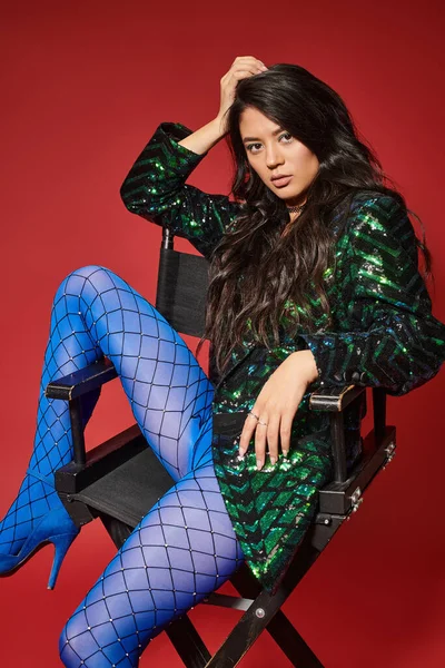 Brunette asian woman in trendy jacket with sequins and blue pantyhose sitting on chair, red backdrop — Stock Photo