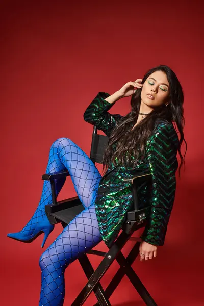 Attractive asian model in jacket with sequins and blue pantyhose sitting on chair, red backdrop — Stock Photo