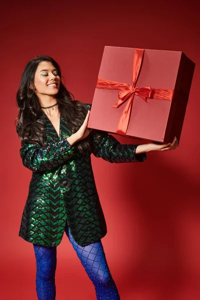 Cheerful asian woman in jacket with sequins and blue pantyhose holding present on red backdrop — Stock Photo