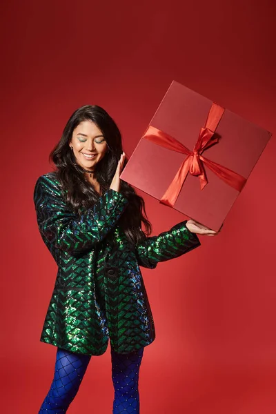 Happy asian woman in jacket with sequins and blue pantyhose holding present on red backdrop — Stock Photo