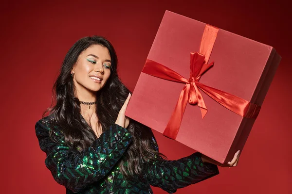 Happy asian woman in green jacket with sequins holding present on red backdrop, Merry Christmas — Stock Photo