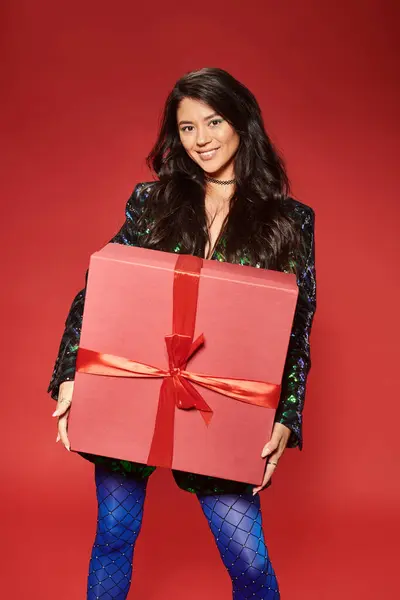 Joyeuse femme asiatique en veste verte avec paillettes tenant boîte cadeau sur fond rouge, Joyeux Noël — Photo de stock