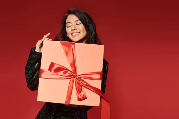 Pleased asian woman in green jacket with sequins holding wrapped Christmas present on red backdrop — Stock Photo