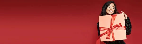 Pleased asian woman in green jacket with sequins holding wrapped Christmas present on red, banner — Stock Photo