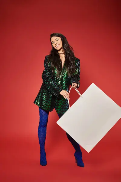 Negro viernes, alegre asiático mujer en verde chaqueta con lentejuelas celebración enorme bolsa de compras en rojo - foto de stock