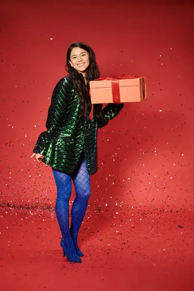 Merry Christmas, cheerful asian woman in green jacket with sequins holding present, festive confetti — Stock Photo