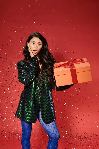 Feliz Natal, chocado mulher asiática em casaco verde com lantejoulas presente de abertura, confete festivo — Fotografia de Stock