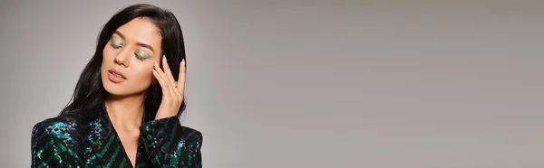 Attractive asian woman in green jacket with sequins and bold eye makeup posing on grey, banner — Stock Photo