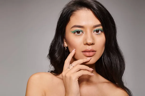 Morena mujer asiática con maquillaje de ojos verdes y hombros desnudos posando sobre fondo gris, mirada audaz - foto de stock