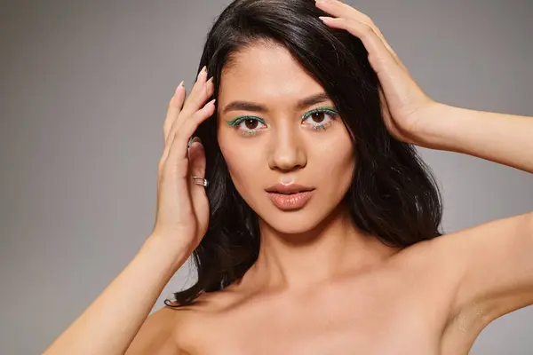 Bruna donna asiatica con scintillante trucco occhi verdi e spalle nude in posa su sfondo grigio — Foto stock