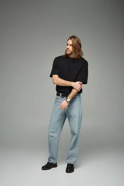 Full length of bearded handsome man in black t-shirt and jeans standing on grey background — Stock Photo