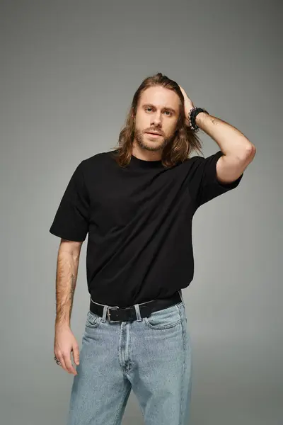 Бородатый красивый мужчина в черной футболке и джинсах, регулирующий длинные волосы и стоя на сером фоне — стоковое фото