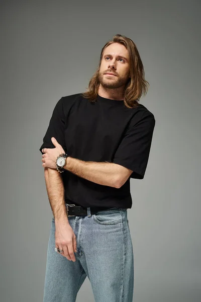 Bearded and handsome man with long hair posing in black t-shirt and jeans on grey background — Stock Photo