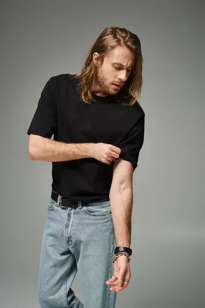 Bearded handsome man with long hair posing in jeans adjusting sleeve on t-shirt on grey background — Stock Photo
