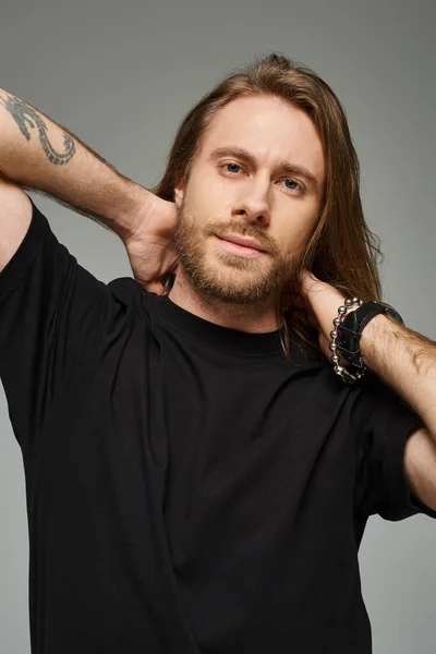 Homem barbudo e bonito com cabelos longos posando em jeans e camiseta com as mãos atrás do pescoço no cinza — Fotografia de Stock