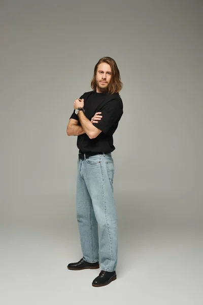 Повна довжина бородатого і красивого чоловіка з довгим волоссям позує в джинсах і футболці на сірому фоні — стокове фото