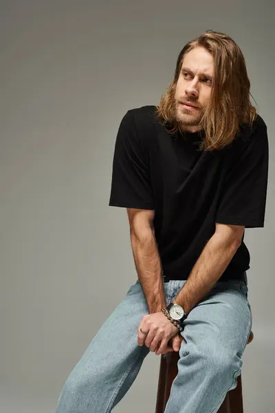 Porträt eines bärtigen und gut aussehenden Mannes mit langen Haaren in Jeans und T-Shirt, der auf einem Stuhl auf grau sitzt — Stockfoto