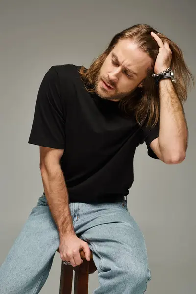 Portrait of bearded and handsome man with long hair in jeans and t-shirt sitting on chair on grey — Stock Photo