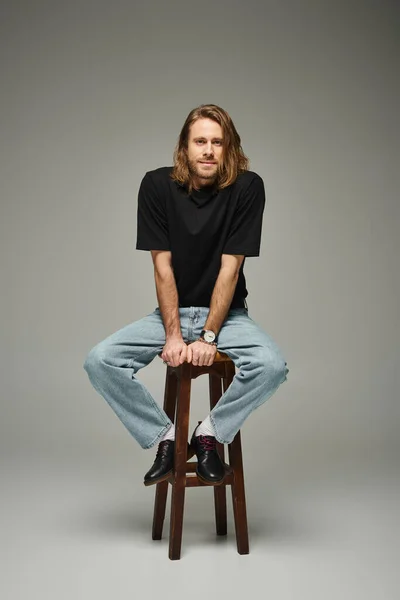 Volle Länge des bärtigen gut aussehenden Mannes mit langen Haaren sitzt in Jeans und T-Shirt auf einem hohen Hocker — Stockfoto