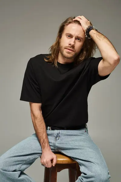 Porträt eines bärtigen und gut aussehenden Mannes mit langen Haaren in Jeans und T-Shirt, der auf einem Stuhl auf grau sitzt — Stockfoto