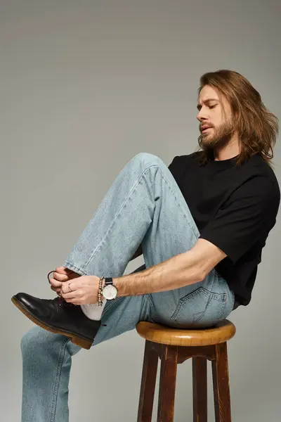 Bearded and handsome man with long hair in jeans and t-shirt sitting on chair and tying shoelaces — Stock Photo