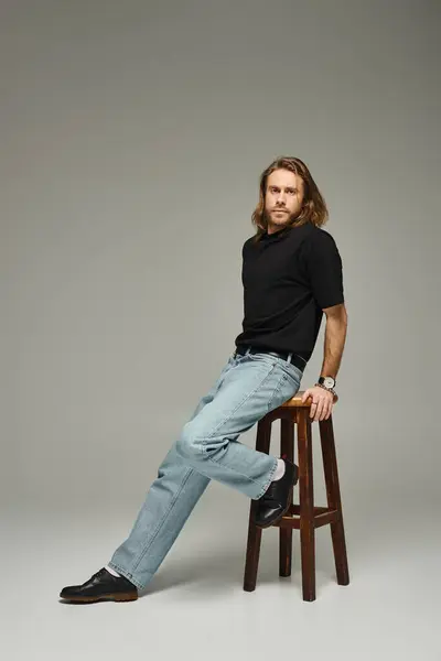 Vollbärtiger Mann mit langen Haaren posiert in Jeans und T-Shirt, während er sich auf einen hohen Hocker lehnt — Stockfoto
