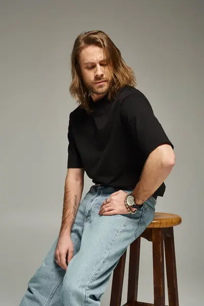 Bärtiger und gutaussehender Mann mit langen Haaren in Jeans und T-Shirt auf Stuhl sitzend mit der Hand an der Hüfte — Stockfoto