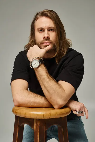 Hombre barbudo y guapo con el pelo largo en jeans y camiseta posando junto a la silla de madera en gris - foto de stock