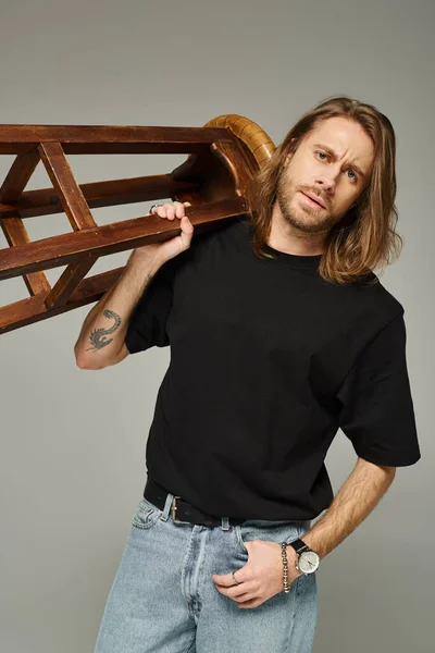 Portrait of bearded man with long hair posing in jeans and t-shirt while carrying high stool — Stock Photo