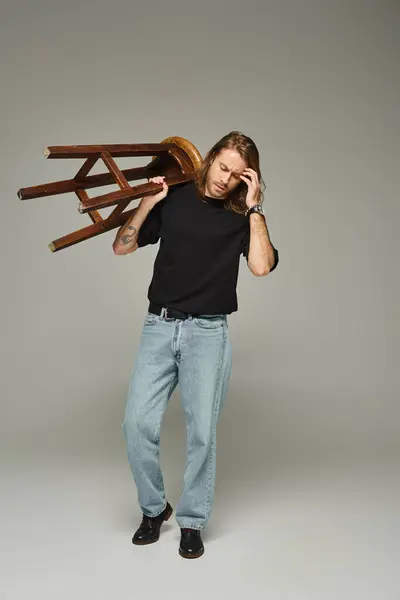 Full length of bearded man with long hair posing in jeans and t-shirt while carrying high stool — Stock Photo