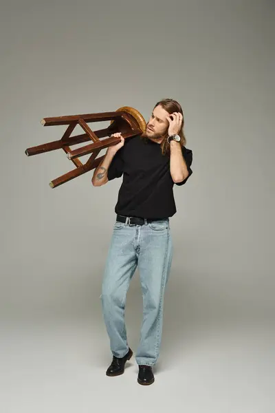 Full length of bearded man with long hair posing in jeans and t-shirt while carrying high stool — Stock Photo