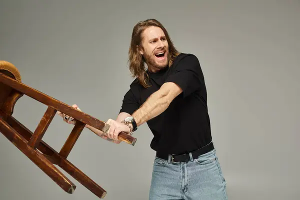 Hombre emocional con el pelo largo en jeans y camiseta llevando taburete alto y gritando en gris - foto de stock